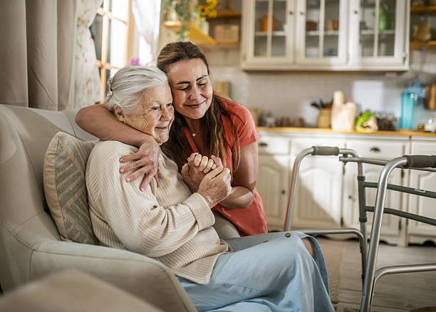 Adult daughter embracing her elderly mother on a couch, with a walker nearby in their home